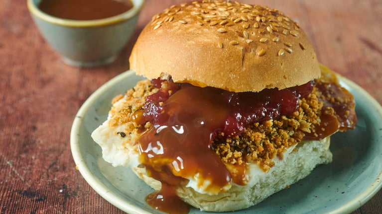 A seeded bap on a plate on a table with turkey, stuffing and gravy and a side pot of more gravy